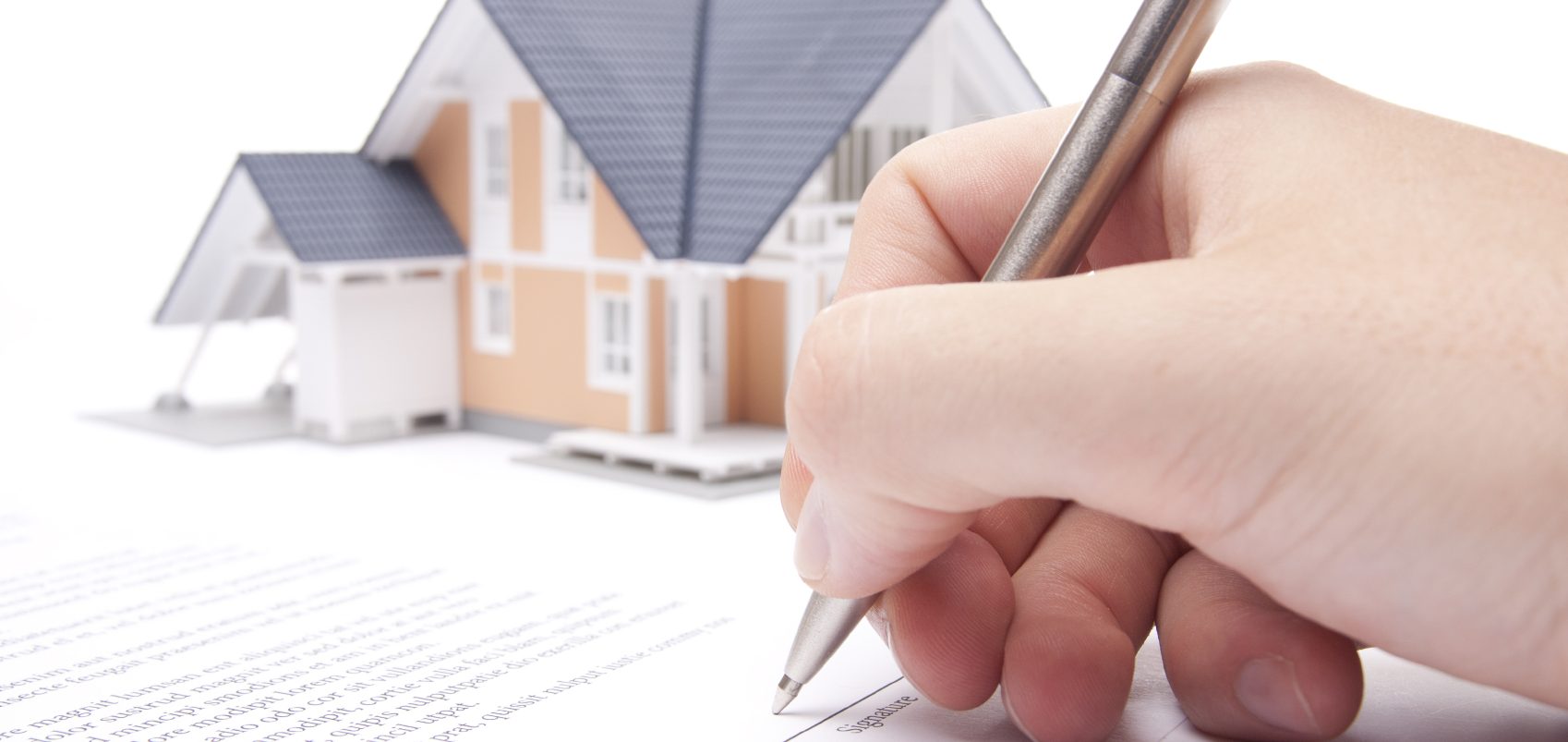 Main dans l'action de signer un document, avec une maison en arrière-plan.
